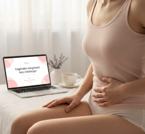 Krupni kadar žene u nežno roze majici koja sedi na krevetu i drži ruku na donjem delu stomaka, dok se u pozadini vidi laptop sa naslovom bloga o vaginalnim simptomima, u svetlom ambijentu