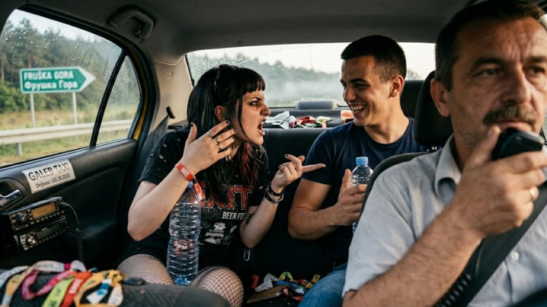 U taksiju koji vozi za Frušku goru: mlada, pijana darkerka viče na nasmejanog mladića pored nje, dok oboje drže flaše piva. U pozadini je ozbiljan taksista koji vozi, a auto je pun haotičnih stvari s Exit festivala.