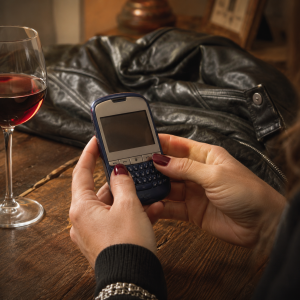 Ženske ruke drže stari mobilni telefon iz ranih 2000-ih na drvenom stolu, pored čaše crnog vina i crne kožne jakne, stvarajući nostalgičnu i emotivnu atmosferu ponovnog kontakta posle mnogo godina.